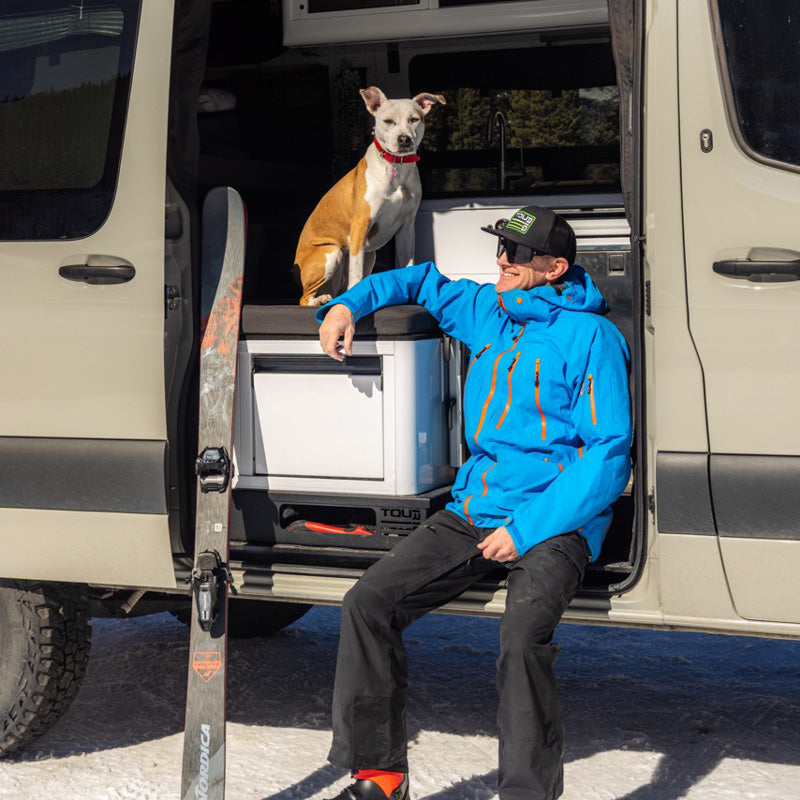 Man in blue ski jacket relaxes by his camper van with a dog sitting inside and skis leaning nearby.