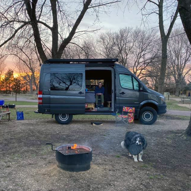 Camper van parked at a wooded campsite during sunset, with a campfire and a dog nearby.