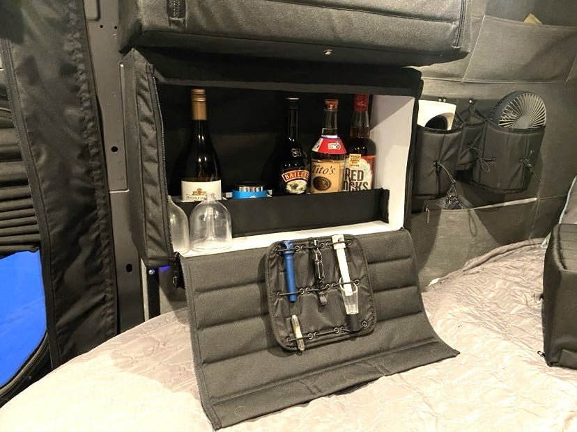 Fold-down camper van bar cabinet stocked with liquor bottles, glasses, and organized bar tools