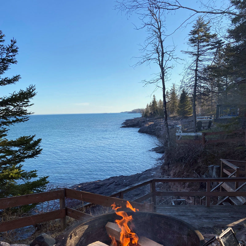 Campfire burns near a rocky lakeshore surrounded by pine trees under a clear blue sky.