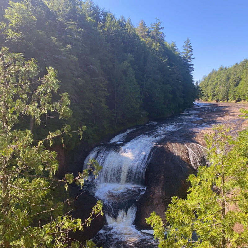 Water cascades over rocky ledges surrounded by lush forest under a clear blue sky.