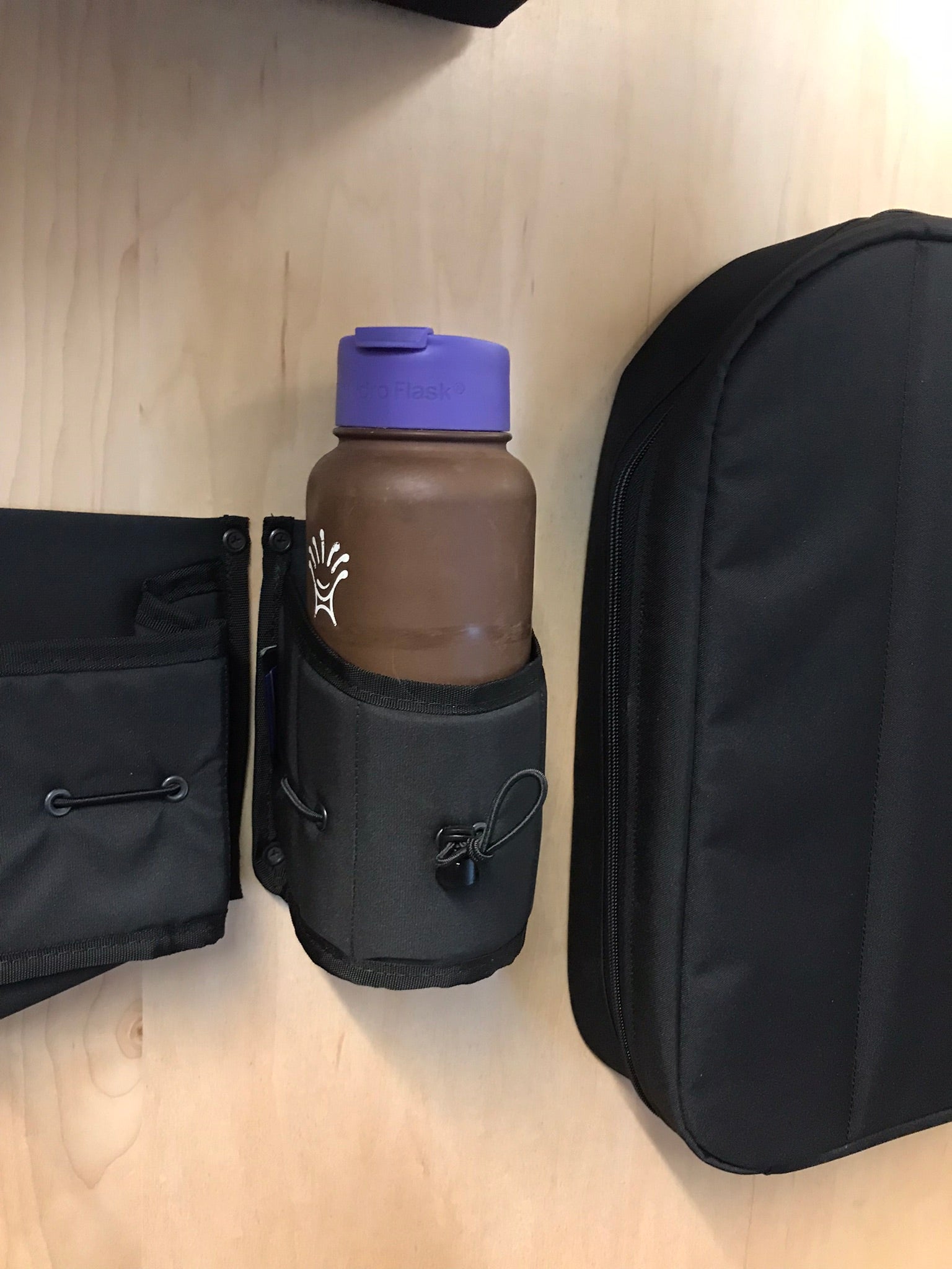 Black fabric water bottle holder securely carrying a Hydro Flask bottle, shown next to other van storage accessories on a wood surface.
