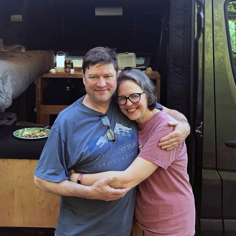 Jeff and Lisa Knight smile and hug in front of their camper van during a road trip.