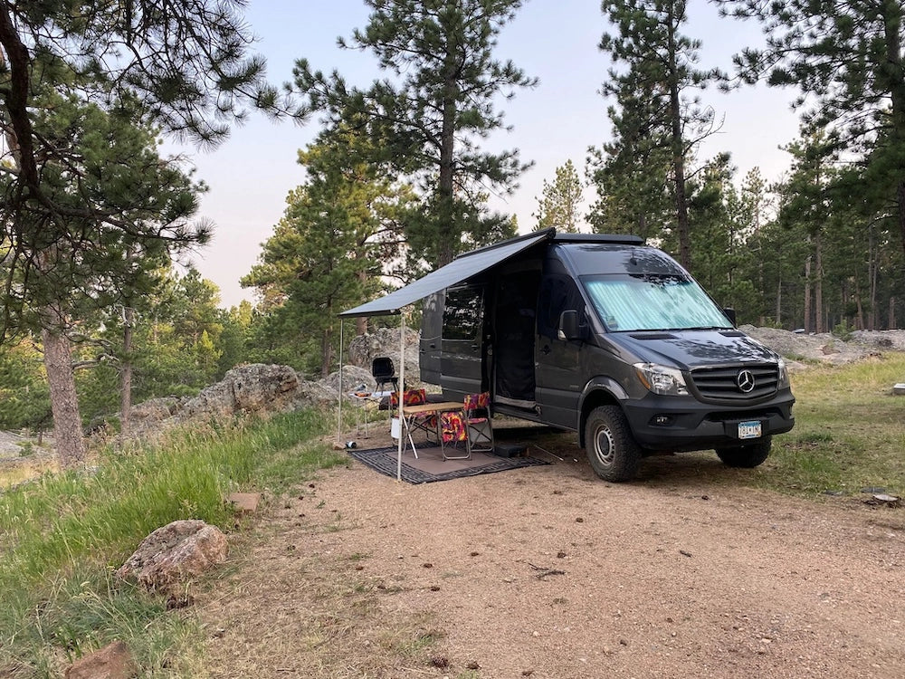 Mercedes-Benz Sprinter camper van parked at a forest campsite with awning extended and chairs set up.