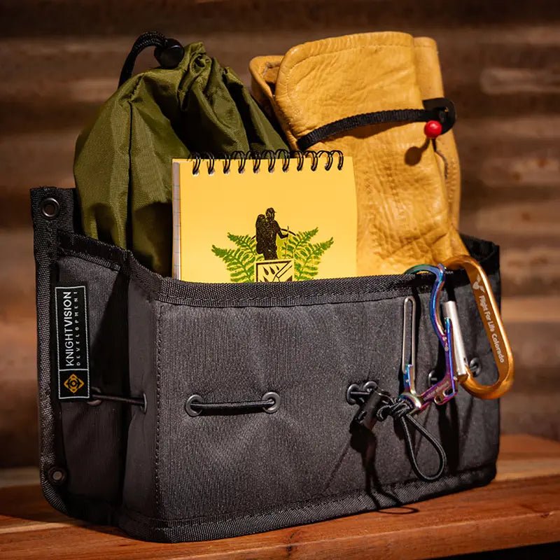 The Spice Cache by Knight Vision Development shown in use, holding a yellow notebook, tan gloves, a green gear sack, and attached carabiners.